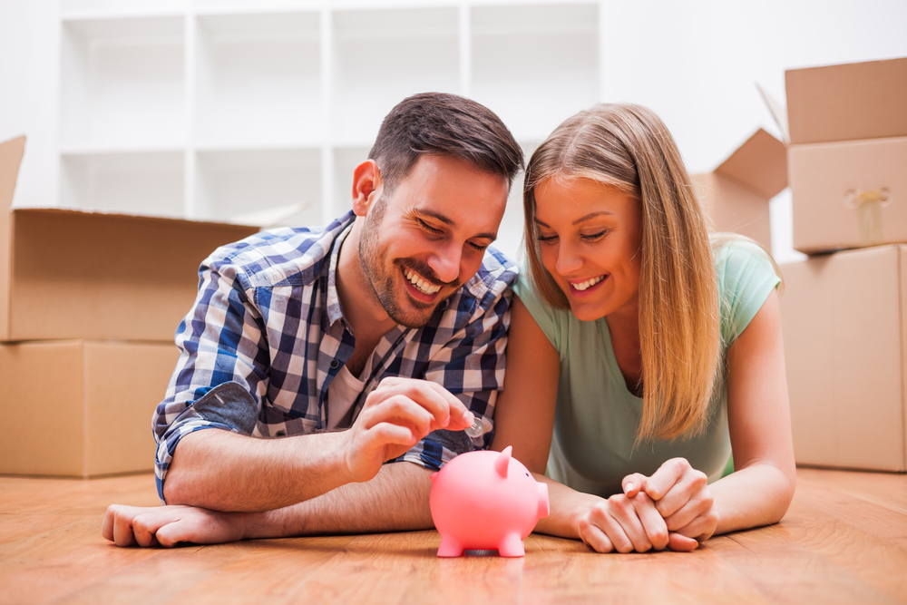 A couple inserting coins into a piggy bank while around moving boxes.