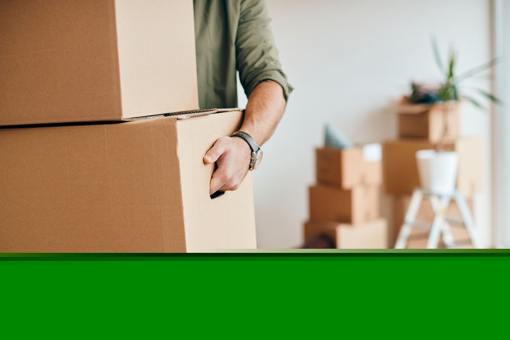 Man carrying two moving boxes with other boxes to move in the background.