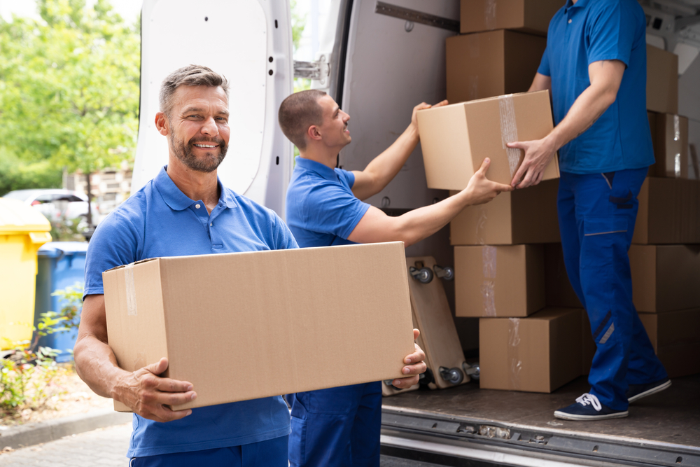 Professional Mover holding a moving box and smiling, while two other movers load boxes into a moving van.